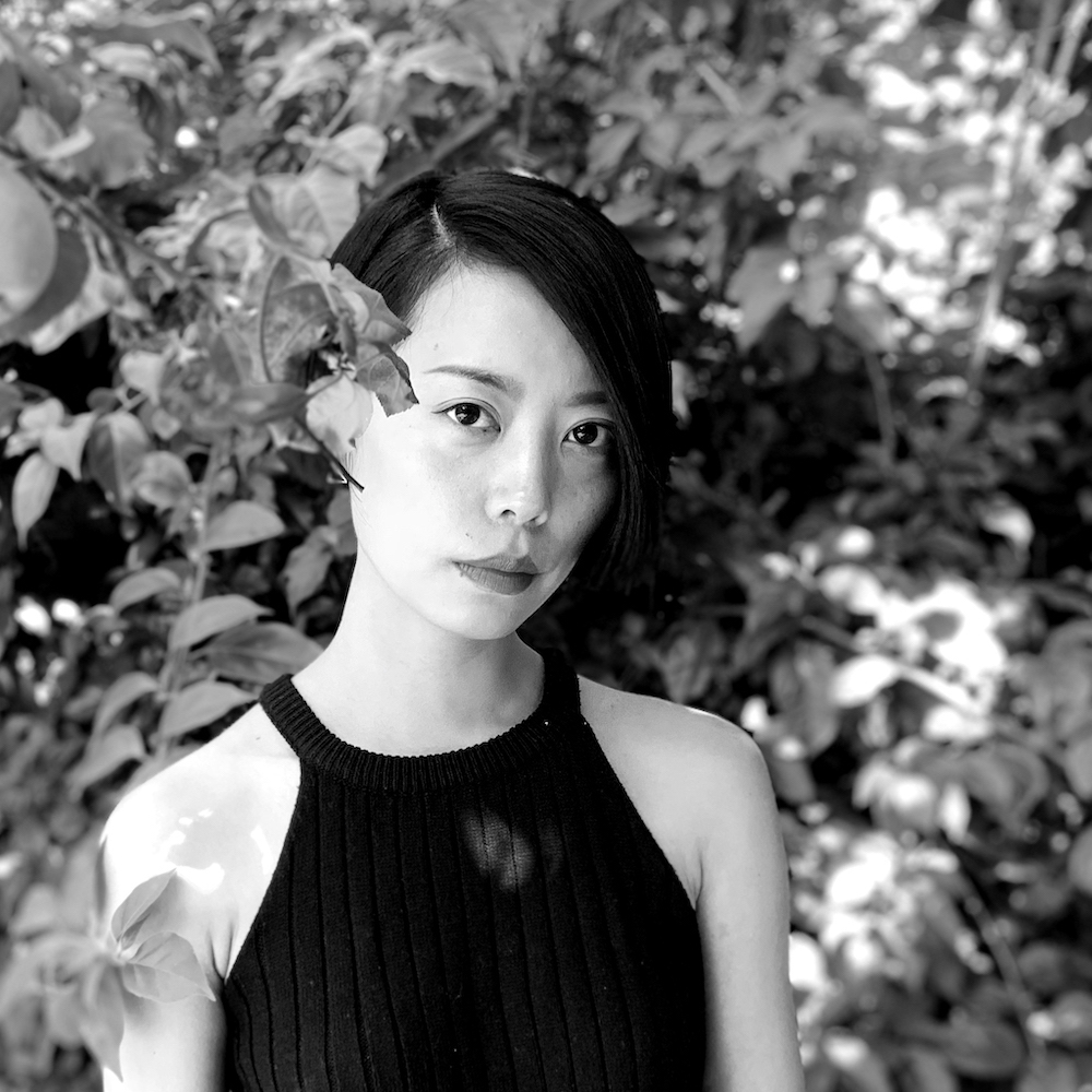 Qianqian, a non-binary Chinese person with black short hair, wearing a black tank top, standing next to an apple tree, with a lush garden in the background.